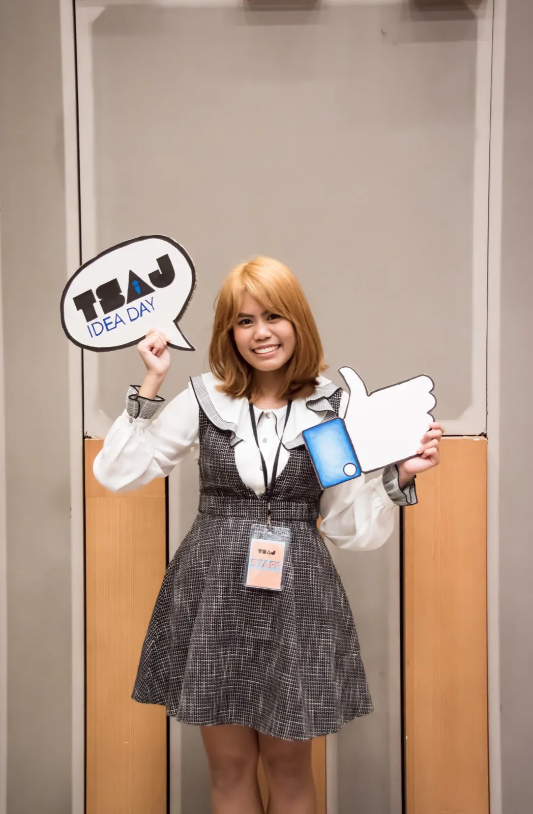 Ornida wearing a staff badge, holding a TSAJ Idea Day speech bubble sign and a thumbs up prop